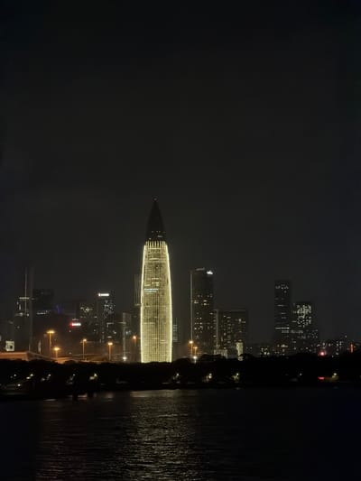 The image captures a nighttime cityscape with a body of water in the foreground, likely a river or bay. The scene is dominated by the illuminated skyline of a modern city. Several tall buildings, featuring numerous lit windows, punctuate the dark sky. A prominent, distinctively shaped skyscraper, appearing conical or pointed, is brilliantly lit with a pattern of horizontal golden lights, standing out against the darker buildings surrounding it. The water in the foreground reflects the city lights, creating a shimmering effect. The overall impression is one of a vibrant, bustling metropolis at night.

The time of day is clearly night, with the darkness of the sky and the prominence of artificial lighting. The weather appears calm, as suggested by the relatively smooth water surface and the clarity of the distant lights. While no people are directly visible, the lit buildings imply human activity and the presence of a populated urban environment. The location is Nanshan District, China, which is known for its modern architecture and waterfront areas.

There are some visible signs of text on the buildings, though they are not clearly legible in the original image. For instance, one of the buildings in the mid-ground appears to have a red sign with white text, and another building has a white sign with dark text. The atmosphere evoked is one of urban energy and modern advancement, with the city lights creating a visually striking display against the night sky. The reflections in the water add a layer of depth and visual interest to the composition.