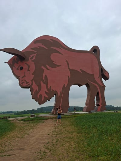 This media file features a large, stylized sculpture of a bull, predominantly reddish-brown with darker brown accents outlining its features, set against an overcast sky. The sculpture is monumental, with its head and horns prominently displayed. The scene appears to be outdoors in a rural or park-like setting, with a field of green grass and crops stretching into the background, bordered by a line of trees. A dirt path runs alongside the sculpture, and a few people are visible near the base of the bull, appearing to interact with the artwork or take in the scenery. One individual is standing closer to the sculpture, and two others are further back, with one appearing to be taking a photo. The overall atmosphere seems casual and leisurely, suggesting a daytime visit to a tourist attraction. The sky is uniformly cloudy, indicating a mild or possibly humid day. The location context is provided as Крошынскі сельскі Савет, Belarus.