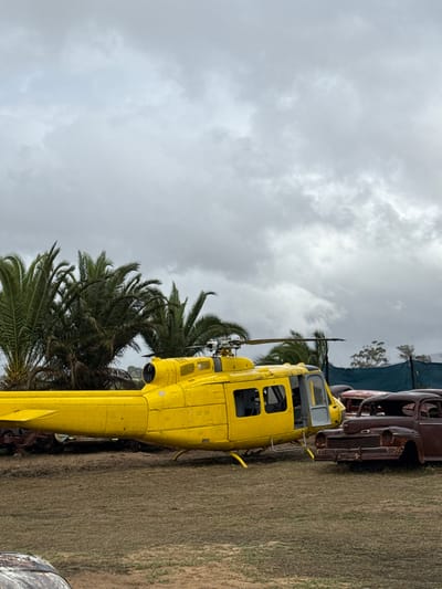 A yellow helicopter rests on the ground near palm trees. Adjacent to the helicopter, several old, rusted cars are visible. The backdrop features an overcast sky and grassy terrain, consistent with an outdoor display of vehicular relics. The location can not be confirmed to be Kraaifontein, South Africa.