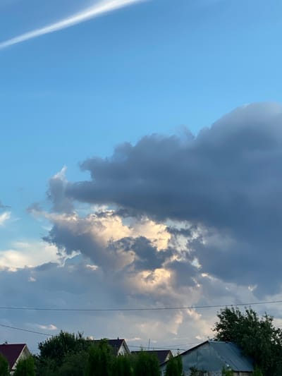 The image depicts a cloudy sky over Oktyabr'skii, Russia. The scene is dominated by a large, dark cloud formation in the center, with patches of lighter, illuminated clouds scattered around it. The sky is mostly blue, suggesting it might be late afternoon or early evening, as indicated by the interplay of light and shadow on the clouds.
In the right portion of the image, parts of buildings and trees are visible. The buildings have dark, sloped roofs, and one appears to be made of brick. Dense green foliage from trees surrounds the structures, hinting at a suburban or rural setting. A thin, white contrail is visible in the upper left quadrant of the image, suggesting an airplane passed overhead recently.
There are no people or animals visible in the image. The overall mood is somewhat dramatic due to the heavy cloud cover, but the presence of the blue sky and illuminated clouds prevents it from appearing entirely somber. No text is visible in the image.