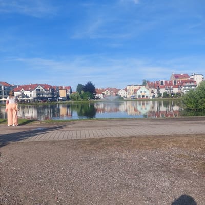 In Mrągowo, Poland, a person stands on a gravel pathway facing a calm body of water. The individual is viewed from behind, wearing an off-white t-shirt and light peach-colored wide-leg pants, with a light-colored tote bag slung over their left shoulder. Their hair is styled with a yellow accessory. Behind the water, a row of buildings with red-tiled roofs lines the opposite bank. A fountain is visible in the water, creating a spray. The sky above is a clear blue with scattered wispy clouds. The foreground consists of a paved walkway made of rectangular grey stones, transitioning to a gravel area. The overall scene depicts a serene waterfront environment in Mrągowo.
