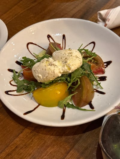 A white, round ceramic plate holds a salad, centrally positioned on a wooden table. The salad consists of two large, oval-shaped portions of soft white cheese, likely burrata, topped with coarse black pepper and a light oil drizzle. Surrounding the cheese are wedges of variously colored tomatoes, including red, reddish-brown, and yellow varieties, intermingled with green arugula leaves. A dark, viscous balsamic reduction or glaze is drizzled in an ornamental pattern across the white plate surface. In the background, to the upper left, a partial view of an additional white dish is visible. To the upper right, a folded, light-colored fabric napkin with thin red stripes is partially visible. The lower right foreground reveals a partial view of a metallic object, likely a utensil. The environment is an indoor dining setting in Chula Vista, United States of America.
