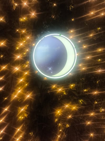 The image depicts a decorative crescent moon illuminated by a neon light, set against a dark background of foliage. The moon appears to be a digital projection or a light fixture, displaying a starry night sky with clouds within its illuminated arc. Numerous small, bright yellow fairy lights are scattered throughout the foliage, creating a bokeh effect and adding a festive atmosphere. The scene is likely part of an outdoor decoration or installation. The location mentioned, Chaidari, Greece, suggests this scene is situated within that city. There are no people or specific actions captured in the image.