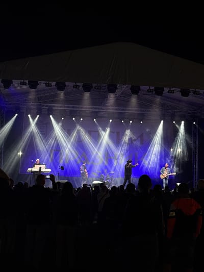 This image depicts a nighttime outdoor music concert in Elin Pelin, Bulgaria. A band is performing on a large stage under a dark canopy supported by metal trussing. The stage backdrop is a blue banner featuring stylized white text, possibly "ЕЛИН ПЕЛИН", and a crest emblem on the left. Bright white spotlights emit distinct beams of light across the stage, which is also illuminated by blue ambient lighting.

From left to right on stage, performers include:
1.  A male individual with light-colored hair, wearing a dark top, seated and playing a white keyboard.
2.  A male vocalist, wearing a dark hat and dark clothing, standing centrally, holding a microphone, with his right arm raised.
3.  A male guitarist, wearing a denim jacket, patterned shirt, and jeans, standing on the right side of the stage, playing an electric guitar.
4.  A drum kit is partially visible behind the central performers.

In the foreground and midground, a large crowd of people, mostly silhouetted against the stage lights, faces the performers. Several individuals are holding up smartphones, presumably recording the event. One person in the right foreground wears an orange and black hooded jacket. The upper portion of the image shows the dark night sky above the stage structure.