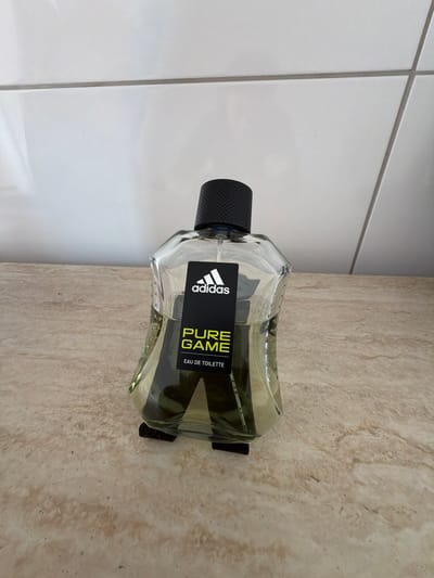 The image shows an Adidas Pure Game Eau de Toilette bottle. The bottle is made of clear glass and contains a light green liquid. It has a black cap and a black label with white and neon green text. The label displays the Adidas logo, "PURE GAME," and "EAU DE TOILETTE." The bottle is placed on a light-colored, textured surface, possibly a countertop or floor, next to a white tiled wall. The lighting appears to be artificial, casting some shadows. There is no person or activity depicted in the image. The overall scene is a still life of a cosmetic product in a home or retail setting. The location is identified as Dorsten, Germany.