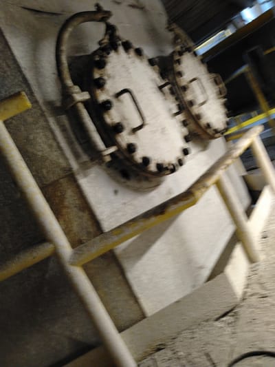 The scene is an interior view of an industrial facility located in Devnya, Bulgaria, presented with a diagonal orientation from the lower left to the upper right. In the central and upper sections, two substantial, circular metal hatches are prominently featured, each secured by multiple dark bolts along its circumference. The left hatch, more fully visible, has a curved metal pipe or conduit attached to its top, originating from the upper-left corner of the frame. The hatches are mounted on a light-colored, possibly concrete or painted, vertical surface. Yellow-painted metal safety railings or handrails, exhibiting wear, dirt, and some paint loss, are visible across the lower and right portions of the image, suggesting a walkway or platform. The immediate surroundings include rough, light-toned industrial surfaces, likely concrete, forming walls and a textured floor in the lower right. The upper background is dim, showing indistinct industrial overhead structures and ambient artificial lighting. No persons are present, and no active interactions or specific actions are occurring.