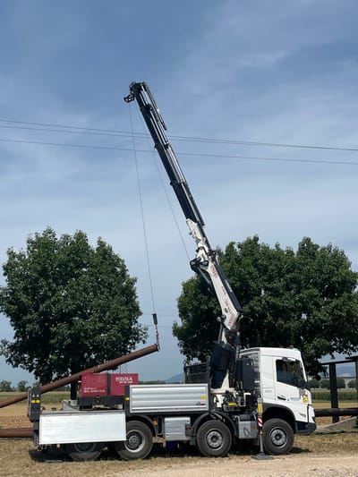 In Castiglione delle Stiviere, Italy, a white truck equipped with a crane is actively engaged in lifting a long, rusty metal pipe. The pipe is suspended from the crane's hook, positioned diagonally across the frame, and appears to be in the process of being loaded onto the truck's flatbed. Beside the truck's rear, a red generator with "MOSA SUPER SILENCED" branding is visible. The truck itself has a silver flatbed and various mechanical components associated with the crane. The scene is set outdoors under a partly cloudy blue sky, with lush green trees framing the background. Power lines are also present, stretching horizontally across the sky. The ground is a mix of dirt and gravel. No people are visible in the image.
