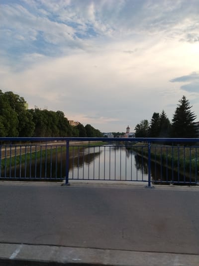 The image depicts a view from a bridge overlooking a river in the city of Strakonice, Czechia. The perspective is from ground level, looking forward and slightly downward towards the river. A blue metal railing is prominently featured in the foreground, with the river flowing beneath it. On the far bank of the river, lush green trees line the shore, with buildings visible behind them. In the distance, a clock tower with a red roof stands out against the sky. The sky itself is a mix of blue and white clouds, suggesting it is either early morning or late afternoon. The water in the river is calm and reflects the surrounding scenery. The road surface of the bridge is visible in the lower portion of the image. The scene is serene and captures a typical urban landscape with natural elements.