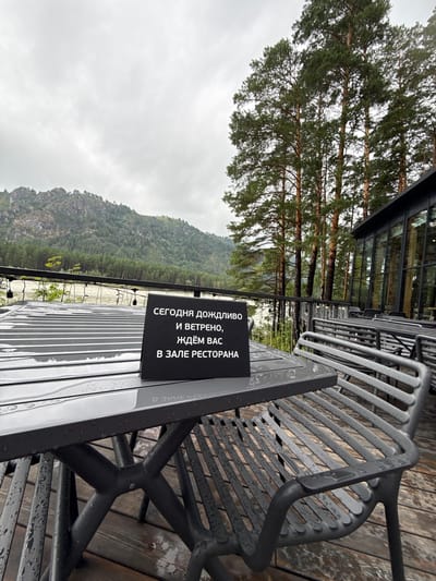 An outdoor seating area with tables and chairs is set up on a wooden deck overlooking a wide river and forested mountains under a cloudy sky. A black sign on a table reads in Russian: "Today it is rainy and windy, we are waiting for you in the restaurant hall." The tables and chairs are wet from rain, and water droplets are visible on the surfaces. The scene suggests a restaurant or cafe with an outdoor seating option that is currently not in use due to the weather.
