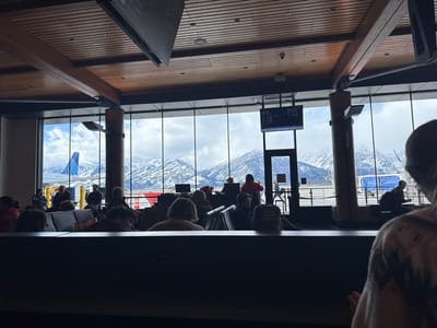 An airport gate area filled with people, viewed from within. A large window reveals a snowy mountain range outside. A United Airlines plane is visible on the tarmac. A digital flight information display board hangs above a doorway. Seating lines the foreground, partially obscuring the view. People are seated, standing, or walking around the gate area. The ceiling is high, with wooden beams and modern lighting fixtures.