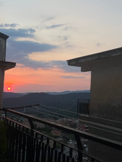 This is a photograph taken in Vallo della Lucania, Italy, likely during sunset, given the warm hues of orange and pink in the sky and the low position of the sun. The image is oriented vertically, with the sunset prominently featured in the center.
The scene is viewed from a high vantage point, possibly a balcony or rooftop, with architectural elements of a building in the foreground. These include concrete structures and metal railings, suggesting an urban or residential setting. Below the sunset, silhouetted mountains form a dramatic backdrop. Further in the distance, a town or village is visible, with red-tiled roofs and buildings characteristic of Italian architecture. The presence of streetlights suggests that dusk is settling in.
The sky is filled with clouds, some dark and moody, others catching the sunset's glow. The overall atmosphere is serene and picturesque, evoking a sense of peace and natural beauty. There is no visible text in the image. The time of day is clearly sunset, and the weather appears clear with some cloud cover, creating a beautiful interplay of light and shadow. The location is hinted at by the architectural style and the mountainous terrain, which aligns with Vallo della Lucania.