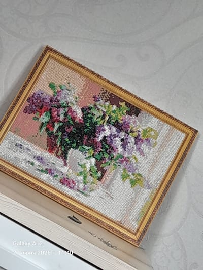 This image shows a framed needlepoint artwork depicting a vase of lilacs on a table, placed against a textured wallpaper background. The artwork is a detailed representation of lilacs in various shades of purple, pink, and green, with a brown vase and a white tablecloth. The frame is ornate and golden. The image also captures a portion of a shelf or surface the artwork is resting on, which appears to be made of wood. In the bottom left corner, a text overlay from a device indicates "Galaxy A12" and "27 июня 2026 г. 11:40" which translates to "June 27, 2026, 11:40". This information, especially the date, is likely metadata from the device used to take the picture and does not relate to the actual content of the artwork or its environment. The specific location in Kara-Köl City, Kyrgyzstan is not visually indicated within the image itself, only through the prompt's context. The lighting suggests an indoor setting, possibly a home.