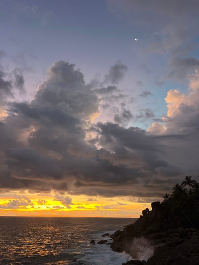 This vertical image, depicting a coastal scene in Rumassala, Sri Lanka, captures a dramatic moment at either sunset or sunrise. The sky dominates the left side, filled with a dynamic array of clouds. These range from dark, brooding grey masses to lighter, more ethereal formations, some tinged with warm orange and pink highlights from the low-lying sun. In the upper left quadrant, a distinct crescent moon is visible against a softer, purplish-blue patch of sky.

To the right, the ocean stretches towards the horizon, spectacularly reflecting the intense golden and orange light of the sun in a wide, luminous band across its surface. The water appears active, with visible ripples and waves, particularly closer to the shore.

In the upper right, a dark, rugged rock formation covered with silhouettes of palm trees juts into the frame. Waves are clearly crashing against the base of these rocks, creating a white splash of foam. The scene evokes a sense of both drama and serene majesty, with the contrasting light and shadow enhancing the natural beauty. There is no visible text in the image.