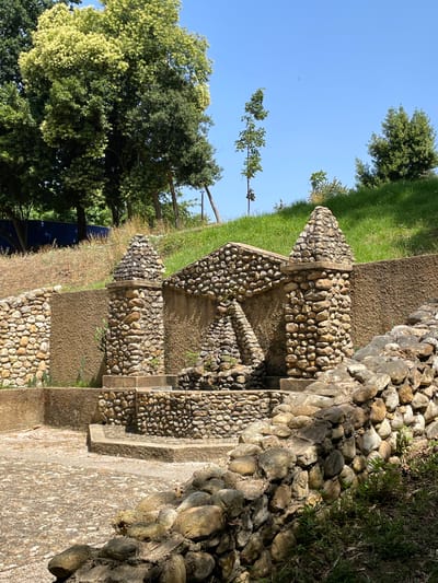 The image depicts a decorative water feature constructed from natural stones. The structure is set within a park-like environment. Lush green grass covers a sloping hillside in the background, with several trees dotting the landscape. The foreground features a paved area made of smaller stones, with a larger stone wall bordering the right side of the frame. The water feature itself is multi-tiered and made of rounded river stones. Two prominent stone pillars with conical tops flank a central arched opening. Sunlight illuminates the scene, casting shadows and highlighting the textures of the stone and foliage. The presence of a blue tarp in the far left background suggests ongoing construction or maintenance in the area. This scene is located in the Pateo de Saltos, Águeda, Portugal.