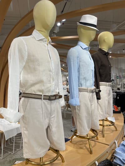 This image displays three mannequins dressed in men's linen clothing, presented in a retail store setting. The mannequins are arranged in a row, facing the viewer from the side. The first mannequin on the left wears a white long-sleeved linen shirt and light beige linen shorts, cinched with a brown and beige braided belt. The second mannequin wears a light blue long-sleeved linen shirt and similar light beige linen shorts, accessorized with a patterned grey belt and a white fedora. The third mannequin is dressed in a dark brown long-sleeved linen shirt and light beige linen shorts, with a patterned grey belt. The mannequins stand on circular, gold-colored stands.

The background reveals elements of a store interior, including wooden beams, track lighting, and shelving units displaying clothing. The overall aesthetic suggests a fashion boutique or department store. The lighting is bright and artificial.

The location of this image is the Alcott store in Marcianise, Italy. This is indicated by visible branding on a displayed belt in the lower right portion of the image. The store appears to be a retail environment focused on apparel. There are no visible people directly interacting with the mannequins; however, a person is partially visible in the background on the left, appearing to be a shopper. The image captures a curated display of summer clothing.