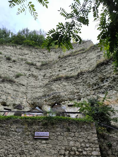 This image depicts a detailed view of a monastic complex built into a natural cliff face in Horaivka, Ukraine. The scene is dominated by a towering, light-colored cliff composed of layered rock, exhibiting erosion and sparse green vegetation, including grasses and small shrubs, growing directly on its surface.
At the base of the cliff, several structures are visible. These include small, simple buildings with sloped roofs, seemingly integrated into natural caves or carved directly into the rock face. One notable structure has a brown-pitched roof with a dark, rectangular opening beneath it. To its right, another dark, rectangular opening is present in the rock. Horizontal wooden or metal beams or railings extend along the front of these structures, indicating a possible walkway or supporting elements.
In the foreground, a robust stone wall, constructed from irregularly shaped, light-colored stones, serves as a retaining structure or boundary. Above this wall, a line of dense green foliage runs horizontally, possibly on top of a railing or fence that is partially obscured. Affixed to the center of this stone wall is a white rectangular sign with dark Cyrillic text that reads: "Душа не може довго Собирать до многово Бог." (The soul cannot long gather to the many God.)
The top portion of the image shows a bright, overcast sky with some visible clouds. Overhanging branches with green leaves are present in the upper left and right corners, partially obscuring the sky and upper cliff edge. Denser green trees and bushes are visible along the top edge of the cliff. No individuals are present in the scene.