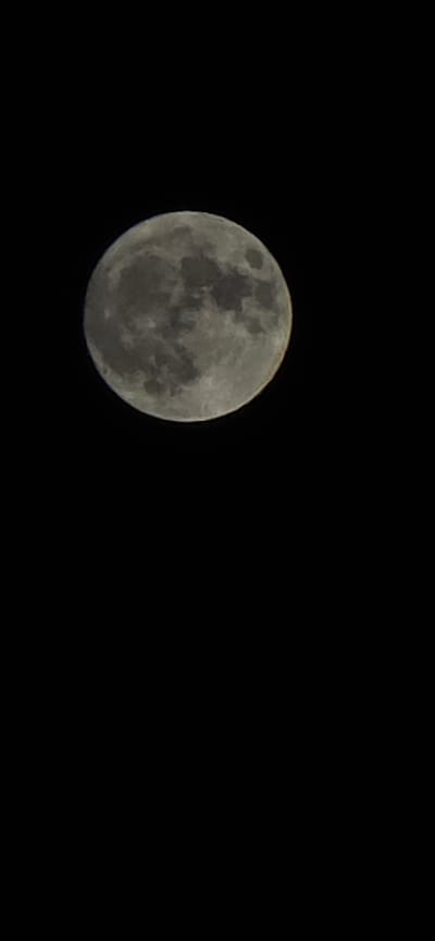 The image shows a full moon against a black sky. The moon is round and grey with visible craters and maria. The lower portion of the moon is illuminated, with the upper portion slightly shadowed. There is a thin, faint reddish-orange glow at the very edge of the moon, possibly indicating an atmospheric effect or a slight overexposure on one side. The rest of the image is entirely black, indicating a very dark environment, likely nighttime. No geographical features or structures are visible, so the location in East Honolulu, United States of America cannot be determined from the image itself.