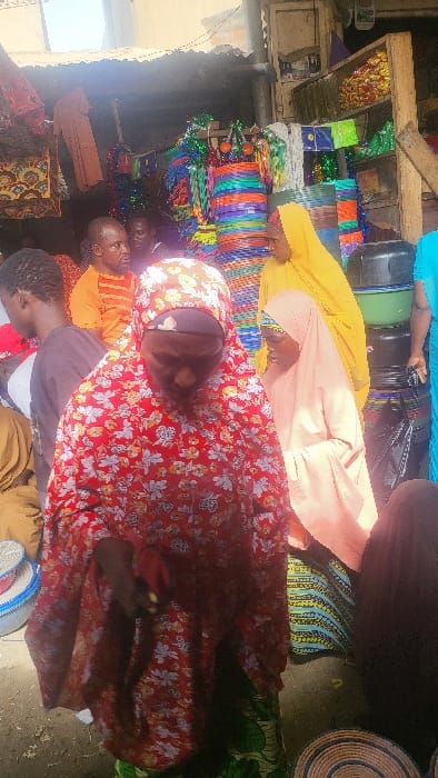 The image depicts an outdoor market scene in Fagge D1, Nigeria, under bright daylight conditions. The ground surface is unpaved earth.
In the immediate foreground, a person, dressed in a red garment with a white and yellow floral pattern and a matching hijab, walks with their head slightly bowed, looking downwards. Their left hand holds a small red object. Beneath the red garment, a green skirt with a yellow and black pattern is visible.
In the mid-ground, multiple individuals are present. To the left of the foreground person, the back and left side of a person with dark hair and a dark top are visible. Behind them, a man with dark skin, wearing an orange striped shirt, is seen from the chest up, looking towards the right. Another partially obscured figure stands behind him. To the right of the foreground person, a woman wearing a bright yellow hijab and dress is visible from the side, facing left. Adjacent to her, another woman wears a light peach-colored hijab and dress, also facing left. Further to the right, a person in a light blue garment is partially visible.
The background features market stalls constructed with corrugated metal roofing and wooden support beams. Various merchandise is displayed. Prominently, colorful plastic mats or baskets, striped in multiple hues including blue, green, orange, and purple, are stacked vertically and suspended from overhead structures. Other hanging items include patterned bags and decorative tassels. On the right side, black and green plastic containers or buckets are stacked. Shelves in the upper background hold packaged goods. On the ground in the lower right, several circular woven mats with concentric patterns are visible.