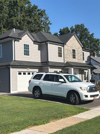 A white Toyota Sequoia is parked in the driveway of a two-story house in Levittown, United States of America. The house has light gray shingle siding and accents of stone on the upper level. A white garage door is visible to the left of the house. The front yard is covered in green grass, and a concrete sidewalk runs along the front of the property. Mature trees with green foliage are visible behind the house, and the sky is a clear blue.