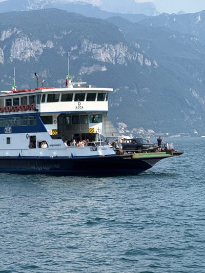 This media file features a ferry boat named "ADDA" with people and cars onboard. The ferry is on a body of water, likely a lake, with a mountainous landscape in the background. The mountains are covered in lush green trees, and some rocky outcrops are visible. The water is a vibrant blue and appears to be slightly choppy, suggesting a breezy day.
On the ferry, several people are visible, some standing and others seated, enjoying the ride. One black car is parked on the deck. The ferry itself is white with blue accents and red railings. The text "ADDA" is prominently displayed on the side of the ferry. Another visible text is "navigazionelaghi.it". The time of day appears to be daytime, with bright sunlight casting shadows, indicating clear weather. The scene evokes a sense of leisure and travel, likely a common mode of transportation for tourists in the region. The location is Loveno, Italy, a place known for its scenic beauty and lake resorts.
