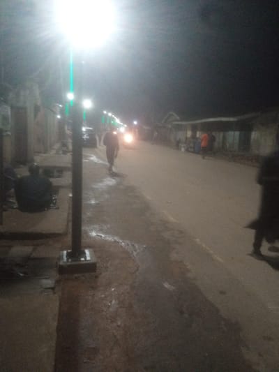 The image depicts a nighttime street scene in Tundun Wada West, Nigeria. A bright street light, with a green luminescence, illuminates the foreground and casts a strong glare. To the left, a person is seated on a concrete structure, facing away from the camera. Further down the street, a person walks away from the camera, dressed in dark clothing. The street is paved and appears wet, with some reflective puddles. Several other figures and structures are visible in the background, including buildings lining the street and what appear to be vehicles. The overall ambiance is that of a dimly lit urban environment at night.