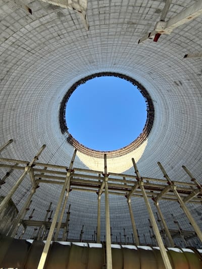 The image presents a striking upward-looking perspective from within a colossal, circular industrial structure, strongly resembling a cooling tower. The vantage point is directly from the base, emphasizing the immense height and scale of the edifice.

The interior walls of the structure are constructed from light grey concrete, characterized by a grid-like or waffle pattern composed of numerous square or rectangular panels. This textured surface appears weathered and somewhat aged, with subtle variations in tone suggesting exposure to elements over time. The cylindrical form of the tower tapers slightly towards the top, perfectly framing a pristine, bright blue sky in a distinct circular opening directly overhead. The clarity of the sky indicates fair weather and daytime.

On the right side of the image, a complex internal framework or scaffolding system is visible, extending vertically. This structure is composed of numerous light-colored beams, possibly made of wood or a similar material, interconnected by horizontal cross-members, forming a multi-tiered, angular pattern. Further to the right and slightly out of focus, darker, rectangular or trapezoidal panels appear to be attached to or supported by this framework. These elements are likely components of the cooling tower's fill material, designed to increase the surface area for water evaporation.

The lighting within the tower is natural, emanating from the circular opening at the top. The direct sunlight highlights portions of the right wall and the internal framework, creating areas of brightness and casting subtle shadows that add depth to the massive structure. There are no people visible in the image, nor is any discernible text present. The overall impression is one of vast industrial architecture, perhaps abandoned or under construction, with a dramatic interplay of concrete, structural elements, and open sky.