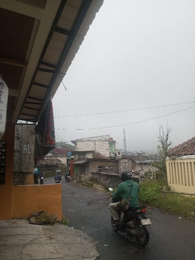 A narrow, paved street winds through a neighborhood on a foggy day. Several buildings, some with tile roofs, line the street. A tall telecommunications tower rises in the distance. Three motorcycles are visible, one of which is moving towards the viewer. Overhanging roof structures appear on the left, with a hanging textile object. The sky is overcast and gray.