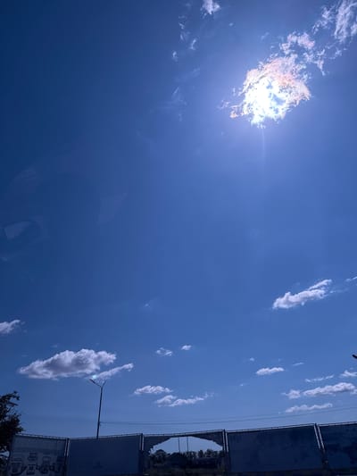 The image captures a bright, sunny sky with scattered white clouds over the city of Прилиманське, Ukraine. The sun is partially obscured by a dense cloud in the upper right quadrant, creating a radiant halo effect and lens flare. In the lower portion of the image, a segmented fence or barrier is visible, with some sections appearing damaged or torn. A tall street lamp stands near the center of the fence, its light fixture partially obscured by a small cloud. To the left of the street lamp, a dark-colored tree is partially visible. The overall scene suggests an outdoor environment with a clear sky and some urban infrastructure.