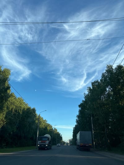 The media file shows a view of a road lined with trees under a bright blue sky with wispy white clouds. The image appears to be taken from a moving vehicle, looking upwards and to the right. In the distance, there are a few vehicles, including a large truck with a gray tarp covering its cargo and a cement mixer truck. The time of day seems to be daytime, likely late morning or early afternoon, given the clear sky and sunlight. The weather appears to be pleasant and fair. The presence of trees and a road suggests a semi-rural or suburban setting. The sky is a prominent feature, with the moon visible as a small, pale crescent. Power lines are visible stretching across the frame, indicating infrastructure in the area. There is no visible text that is clear enough to be read. The overall mood is serene and captures a moment of travel on a clear day.