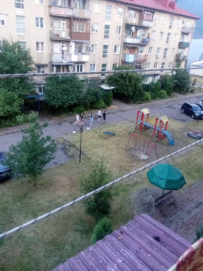 The image shows an outdoor scene in front of a multi-story apartment building, likely in Tiachiv, Ukraine. In the foreground, a clothesline runs across the frame. The middle ground features a paved area with a street, a small playground, and some grassy areas with trees. The playground has a slide, swings, and a merry-go-round. A dark blue car is parked on the street, and a black SUV is parked near the playground.
Several people, appearing to be young adults or teenagers, are walking on the paved area. They are dressed casually and their body language does not convey any strong emotions. A green umbrella is open over a bench in the background. The sky is overcast, suggesting a cloudy or possibly rainy day. The time of day appears to be late afternoon or early evening, given the soft, diffused light. The building itself is old, with visible balconies and satellite dishes, typical of Soviet-era architecture. There is no visible text in the image.
