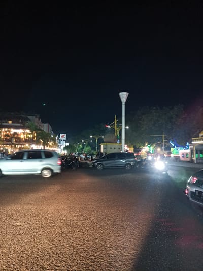 This is a nighttime photograph of a street scene in Bandarbireuen, Indonesia. The image captures a road with several vehicles, including a silver SUV in the foreground on the left, a dark SUV behind it, and a dark car with its taillights illuminated on the far right. The road surface appears to be asphalt with a textured, granular appearance. On the left side of the street, there is a multi-story building with illuminated windows, suggesting it is a commercial establishment, possibly a restaurant or shop, with palm trees in front. Further down the street, a tall, white, decorative lamp post with a unique top structure is visible, along with other street lights and decorative lighting, some of which are colored. There are also numerous parked motorcycles on the right side of the road, near the curb. In the background, trees and more buildings are discernible under the dark night sky. The scene depicts a lively urban environment at night, with artificial lighting illuminating the area.