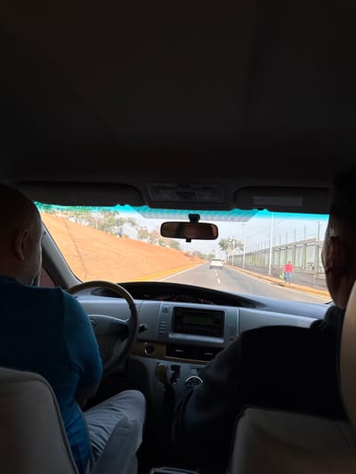 The image shows a car's interior, presumably taken from the back seat. A driver in a blue shirt is visible, along with a passenger in a black shirt in the front seat. The dashboard and steering wheel are visible, along with the rear-view mirror. Outside the car window, a road and a pedestrian walkway with a fence can be seen. A person in a red shirt is on the walkway. The environment suggests a tropical location, and the location is most likely Catia La Mar, Venezuela.