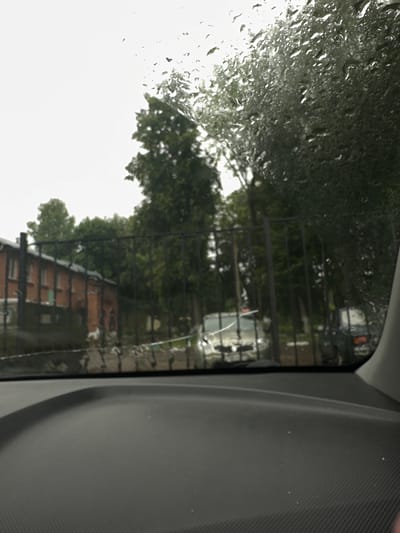 The image shows a view through a rain-streaked car windshield, looking out at a street scene in Monastyrshchina, Russia. The foreground features the textured surface of a car's dashboard. Beyond the windshield, a black metal fence runs across the middle ground. Through the fence, a red brick building with several windows is visible on the left, partially obscured by trees. On the right side of the frame, two cars are parked, one appears to be a light-colored sedan, and the other a darker vehicle. The background consists of dense green trees and an overcast sky. The overall impression is of a damp, grey day.