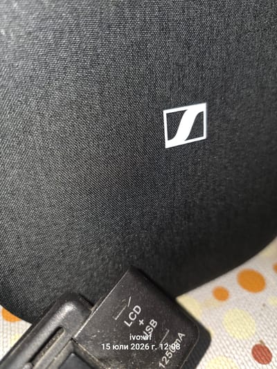The image shows a close-up of a black fabric surface with a white Sennheiser logo on it. In the foreground, a portion of a black electronic device is visible, with text that appears to read "LCD + USB", "ivo.wf", and a date and time "15 юли 2026 г. 12:08". The background has a blurred pattern of orange, yellow, and red dots. The location of Chal, Bulgaria is not visible or inferable from the image.