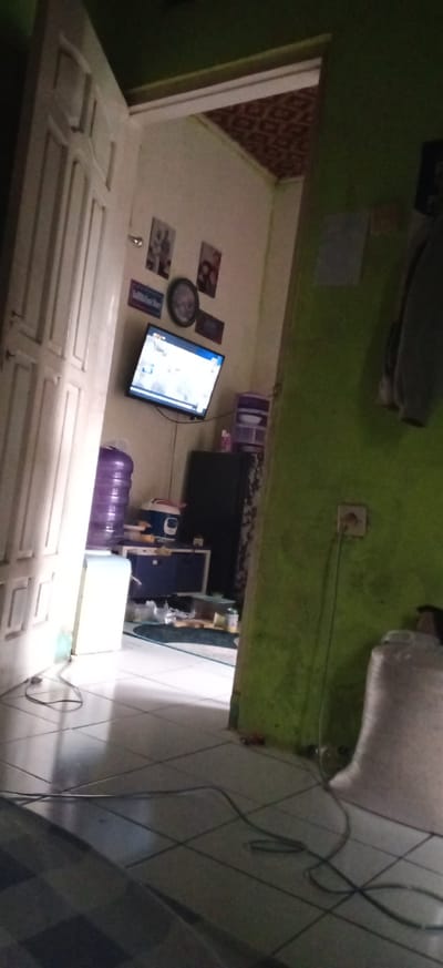 The image presents an interior scene, looking through an open, bright white door into an adjoining room, characteristic of a home in Banjaran, Indonesia. Bright natural light illuminates the foreground, especially the white tiled floor and the left side of the open doorway.
In the room beyond, a flat-screen television is mounted on a light-colored wall, displaying a broadcast that appears to be news or general programming, though specific text on the screen is illegible. The wall around the television is decorated with several framed pictures and a dark, round wall clock. Below the TV, a cabinet or table holds a blue container and a white and blue rice cooker. To the left, a water dispenser topped with a large, purple water bottle is visible. To the right of the water dispenser, a dark refrigerator or tall cabinet stands, with purple plastic storage drawers stacked above it.
The immediate foreground features a vibrant green wall on the right, where an electrical outlet has a cord plugged in, stretching down towards the floor. Various electrical wires are loosely laid across the white tiled floor. In the upper right corner, some clothes or fabric are hanging. The ceiling of the background room displays a distinct patterned design. The bright lighting suggests it is daytime. No individuals are present, giving the impression of a typical, lived-in household space.