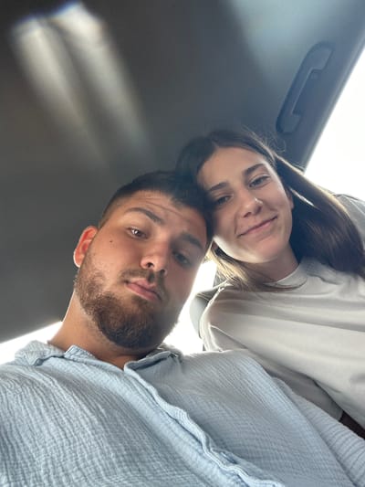 In Dabnik, Bulgaria, a selfie captures two individuals inside a vehicle. A young man with short dark hair and a beard is on the left, wearing a light blue, textured button-up shirt. To his right, a young woman with long dark hair and a light grey t-shirt smiles towards the camera. The car's interior is visible, with a grey headliner and a grab handle. Sunlight streams in from the right, creating bright highlights and casting diagonal shadows across the scene. The perspective is from within the vehicle, looking upwards towards the individuals and the car's ceiling.