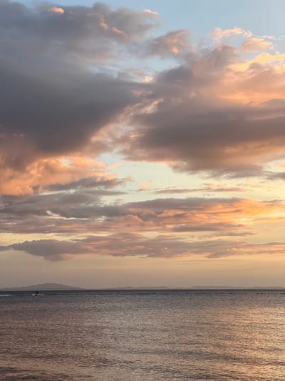 This is a photograph of a sunset over Lake Nicaragua, viewed from Altagracia, Nicaragua. The sky is filled with dramatic clouds in shades of pink, orange, and gray, illuminated by the setting sun. The lake's surface is calm, with gentle ripples reflecting the colors of the sky. In the distance, the silhouette of a shoreline with mountains can be seen. A solitary figure is visible on the water, appearing to be waterskiing or wakeboarding. The overall impression is one of serene natural beauty.