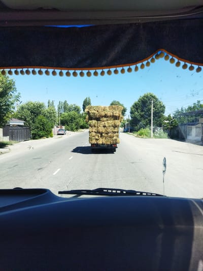 The image, taken from the interior of a vehicle, shows a street scene in Ivanovka, Kyrgyzstan. A truck loaded with bales of hay is traveling down the road ahead. The truck has a license plate that reads "09 021 CP". On the left side of the road, there is a fence and a building. On the right side of the road, there are trees and houses. The sky is clear and blue. The interior of the vehicle is visible in the foreground, including the dashboard and windshield. A decorative trim with yellow pom-poms hangs from the top of the windshield.