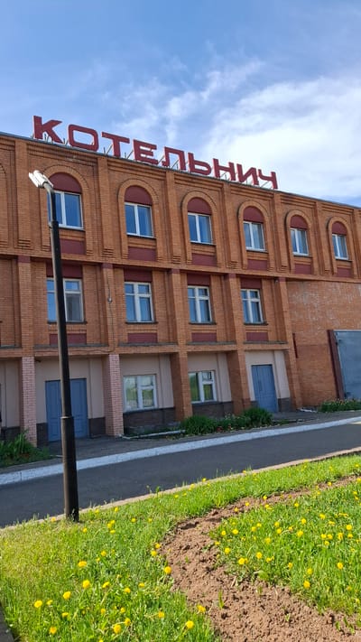 The image shows a multi-story brick building under a partly cloudy blue sky. The building has arched windows on the top floor and a red sign, reading "КОТЕЛЬНИЧ" vertically. Adjacent to the building is a narrow sidewalk and a grass patch with dandelions. The scene appears to be taken in Kotelnich, Russia, based on the sign. There are no visible people, actions, or interactions in the image. A lamp post is present in the foreground.