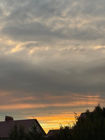 The image depicts a cloudy sky at sunset over a residential area in Uzukovo, Russia. The sky is predominantly gray, with streaks of orange and yellow light visible on the right side, indicating the setting sun. The clouds are textured and appear to be layered, with some areas brighter where the sunlight is breaking through.

In the foreground, on the right side of the image, parts of two houses are visible. The roofs are covered in dark shingles, and the upper portions of the walls are visible, featuring a light-colored siding. A dark green tree with dense foliage partially obscures the houses and the sky. An antenna is visible on one of the house roofs.

The time of day is clearly sunset, given the lighting conditions and the colors in the sky. The weather appears to be partly cloudy, with a dramatic contrast between the darker, overcast portions of the sky and the brightly lit areas where the sun is setting. The scene evokes a sense of calm and the natural beauty of a sunset. No text is visible in the image.