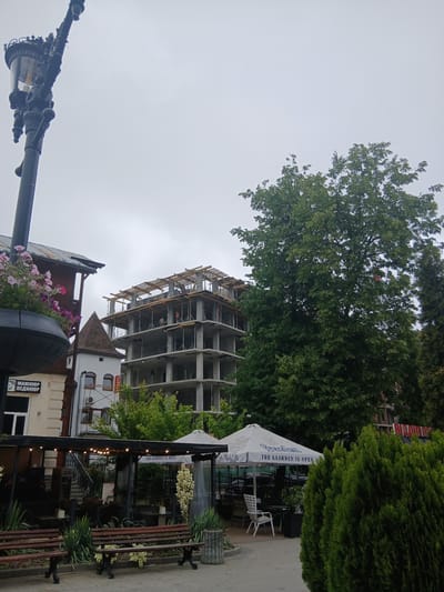 This media file depicts an outdoor scene in Truskavets, Ukraine. The primary subjects are a partially constructed multi-story building, lush green trees, and an outdoor seating area with benches and tables under a large white umbrella that reads "THE GARDEN IS OPEN". The construction site shows scaffolding and workers, indicating ongoing building activity. In the foreground, there is a decorative street lamp and a planter filled with pink and purple flowers.

The scene appears to be during the daytime, with overcast or cloudy weather suggested by the bright, diffused light. The overall atmosphere is one of urban development and outdoor recreation, with the construction juxtaposed against the pleasant setting of the garden area.

Visible text includes "THE GARDEN IS OPEN" on the umbrella. Another sign in Cyrillic text is partially visible on a building in the background, reading "МАНИКЮР ПЕДИКЮР" which translates to "Manicure Pedicure". The presence of these signs, combined with the construction and the outdoor seating, suggests a mixed-use urban environment. There are no overt emotional cues conveyed in the image.