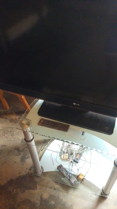 The image shows a TV set on a glass stand in what appears to be a living room in Donga, Nigeria. The TV screen is black, suggesting it is off. Below the TV, on the glass shelf, are several objects including two remote controls, a mobile phone, and a tangle of wires connected to what looks like a power adapter or a small electronic device. The TV stand has metal legs and a shelf made of clear glass. The floor is concrete and shows signs of wear and tear. The lighting suggests it is daytime, but there are no clear indicators of the time of day. The scene is cluttered with electronic devices, hinting at a functional or possibly experimental setup. The overall impression is of a lived-in space with a focus on electronics. The LG logo is visible on the TV frame.