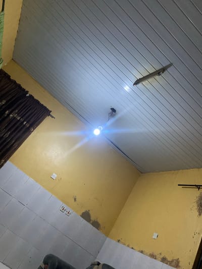 The image presents an indoor room viewed from a low angle, looking upwards towards the ceiling and across the walls and floor. The ceiling is clad in light grey, horizontally paneled material, likely PVC, which reflects the bright illumination. A single, bare light bulb, emitting a strong white light and glare, hangs from the center of the ceiling, serving as the primary light source. Another older, unlit light fixture or housing is attached to the ceiling towards the upper left.
The walls are painted a mustard yellow, showing signs of wear, including patches of discoloration, dampness, or exposed plaster, particularly along the ceiling line and corners. On the right-hand wall, a small white electrical switch or outlet plate is visible. Further down, near the floor, a white double electrical outlet is mounted on the yellow wall. A dark, vertical pipe or conduit runs along the upper right wall.
The floor is partially visible, segmented into white square tiles on the right side and a yellow-painted surface on the left, consistent with the wall color. A dark brown, patterned curtain is drawn across a window or opening, situated along the bottom-center of the frame. Parts of a dark, possibly upholstered object and a green fabric item are visible on the floor at the far right and bottom left, respectively. The strong artificial lighting suggests the scene takes place at night or in a poorly lit environment. This functional room, located in Shen, Nigeria, appears to have modest finishes and some signs of age. No visible text is present.