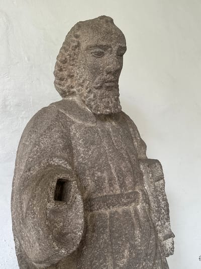 The media file presents a close-up, rotated view of a weathered stone sculpture, likely an effigy, set against a plain, light-colored textured wall. The sculpture is crafted from a coarse, dark grey stone, exhibiting significant erosion and granular texture from age.
The visible portion primarily focuses on the upper body and head of what appears to be a male figure. The head, positioned on the left side of the frame, features a discernible, albeit eroded, face with a carved eye, nose, mouth, and a prominent, textured beard. The body, extending to the right, shows indications of draped clothing or possibly armor, with distinct folds and geometric carvings, including some rectangular shapes.
A notable detail on the sculpture's torso is a prominent square-shaped hole or indentation, surrounded by a swirling or circular depression. This feature suggests it might have once held an inlay, a missing element, or perhaps represents a symbolic carving. The lighting is soft and even, suggesting an indoor display or shaded outdoor environment. No activity is taking place, and no text is visible in the image. The location is Tepotzotlán, Mexico.