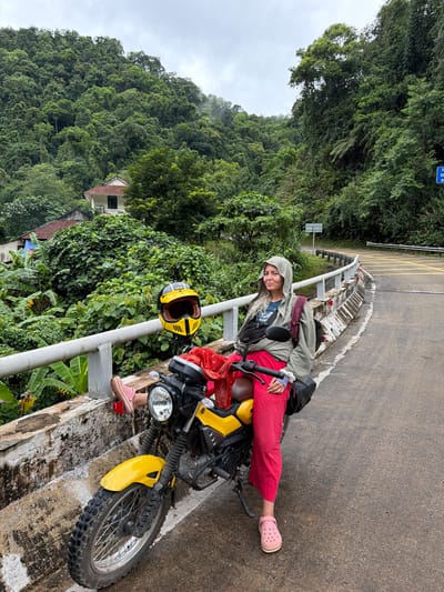 A woman is sitting on a yellow motorcycle parked on the side of a wet, paved road with a concrete barrier and railing. She is wearing a grey hooded jacket, bright pink pants, and pink Crocs. Her left leg is extended over the railing, and a yellow and black helmet rests on the motorcycle's seat. A red cloth is draped over the motorcycle's handlebar. The road curves to the right, marked with yellow lines. In the background, a steep, heavily forested hillside is visible, with scattered houses and lush vegetation including banana trees. The sky is overcast. The scene appears to be in a rural, mountainous area, possibly Thượng Trạch Commune, Vietnam, given the dense jungle and roadside architecture.