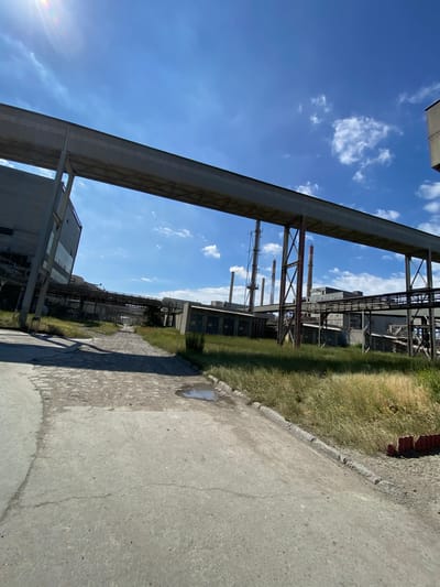 This image depicts an outdoor industrial scene under a bright blue sky with scattered white clouds. A large, elevated conveyor belt system dominates the foreground, stretching across the frame and supported by substantial concrete pillars. To the left, a portion of a large, grey industrial building is visible, angled and showing signs of wear.

The ground in the immediate foreground is a rough, paved surface, showing cracks and a small puddle of water. To the right of the paved area, tall, dry grass grows, bordering a weathered fence.

In the background, several more industrial structures are visible, including buildings and a series of tall smokestacks, some of which appear to be emitting a light plume of smoke. Elevated walkways and pipework connect various parts of the complex. The overall impression is one of an active or recently active industrial facility.

The image is not geographically specific to the city of Police, Poland, and no human presence or activity is discernible.