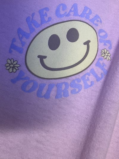 The image shows a close-up of a light purple garment, possibly a t-shirt or sweatshirt, with text and graphics printed on it. The text "TAKE CARE OF YOURSELF" is printed in a darker purple, slightly rounded font, arranged in a circular pattern around a smiley face graphic. The smiley face is light yellow with dark purple eyes and a dark purple curved line for a mouth. Two small, light-colored daisy graphics are placed on either side of the smiley face. The garment appears to be made of a soft, textured fabric. The lighting suggests the photo was taken indoors, with the garment likely displayed in a retail setting or owned by an individual. There is no information in the image to determine its location in Lynnwood, United States.