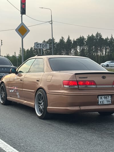 In Solnechnyi, Russia, on a cloudy day, a tan Toyota Mark II Tourer V sedan is stopped at a traffic light. The car is positioned on a paved road, with other vehicles and a dense line of trees visible in the background. A traffic light displays a red signal with a "30" speed limit indicator. A diamond-shaped yield sign and a rectangular "STOP" sign are also present on poles. The car's license plate reads "K 987 XE 186 RUS". The vehicle has aftermarket wheels and several decals on its side. There are no people clearly visible in the immediate vicinity of the car.