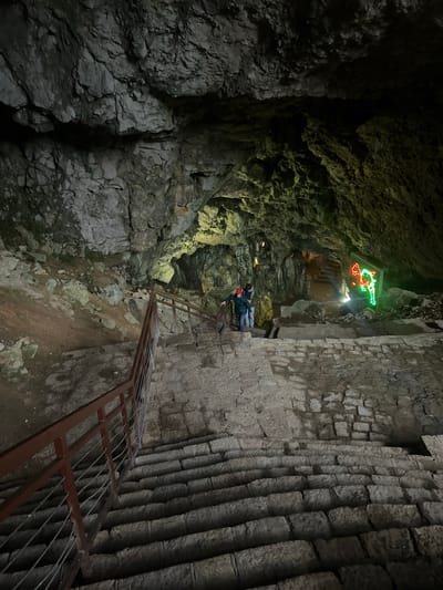 The image depicts the interior of Resavska Pećina (Resava Cave) in Despotovac Municipality, Serbia. The scene is illuminated by artificial lighting, revealing a constructed visitor pathway through the natural cave environment.
In the foreground, a wide, uneven stone staircase descends towards the viewer, equipped with a metal handrail on the left featuring a reddish-brown top bar and multiple horizontal wire cables. The stone steps appear damp or worn.
In the midground, two individuals are visible on a landing area between sets of stairs. One person, wearing a dark jacket and a red cap, stands closer to a secondary railing segment on the left. The second person, largely obscured, stands slightly behind and to their right, wearing a lighter-colored top. Both individuals appear to be descending the pathway. To the left of the pathway, the natural cave floor consists of loose rock fragments and soil.
The cave walls and ceiling throughout the visible area display rugged, irregular rock formations. Artificial lights cast various hues, with some areas exhibiting a yellowish-green tint on the rock surfaces.
In the background, further into the cavern, a prominent display board with red and green neon lights outlines a map or schematic, characteristic of the Resava Cave's visitor routes. Beyond this, a darker passage suggests additional chambers or pathways. The overall environment is a semi-dark, subterranean space made accessible and traversable by engineered stone steps and railings.