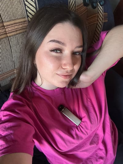 A young woman lies on a striped couch, her head turned towards the camera. She has dark hair and is wearing a bright pink t-shirt. A silver harmonica is clipped to the t-shirt on her chest. The lighting suggests it is daytime, and the scene appears to be indoors, possibly in a home in Krasnokamsk, Russia. The woman's expression is relaxed and she appears to be posing for the photo. There is no visible text in the image.